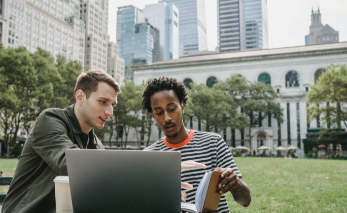 two men looking at notebook in front of silver laptop outside in a park.