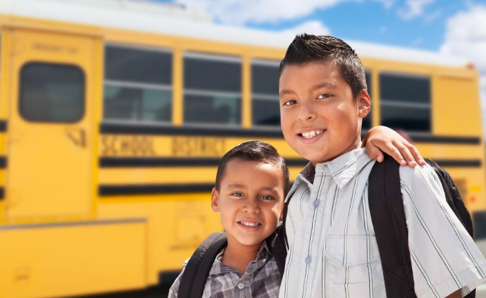 Two children wearing backpacks standing next to a school bus.