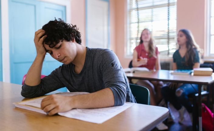 Stressed teen boy studying at desk in classroom.