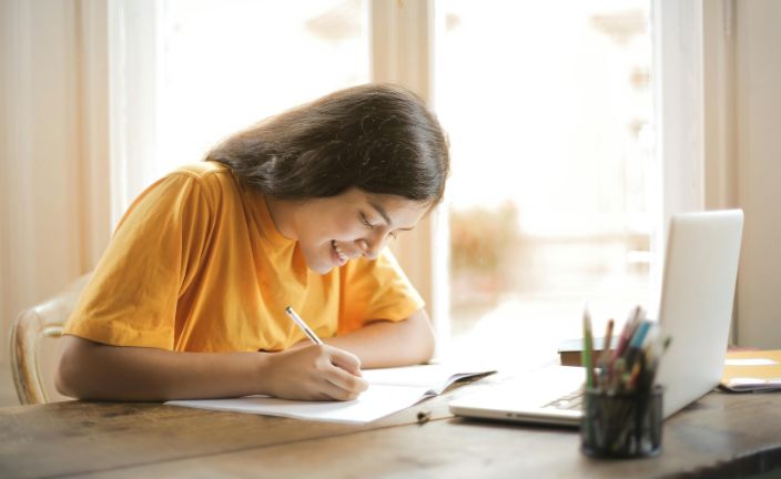 Girl in yellow shirt taking notes in front of laptop.