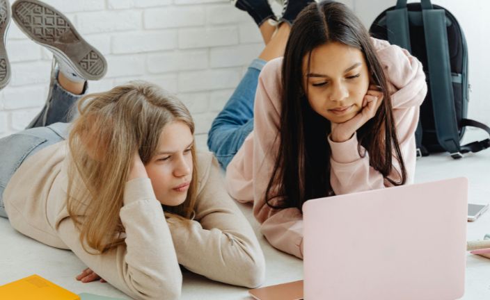 Teen girls studying on laptop in school hallway.